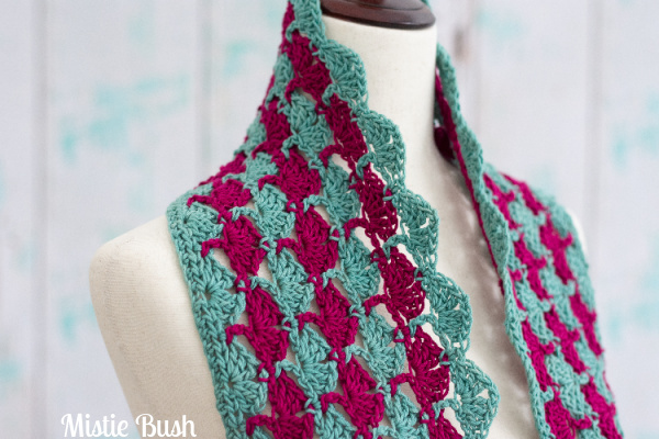 A close-up of the Magade Cowl, a crocheted accessory featuring alternating vertical stripes of jade green and magenta. The cowl displays a lacy, textured stitch pattern and is draped around the neck of a neutral-colored dress form.