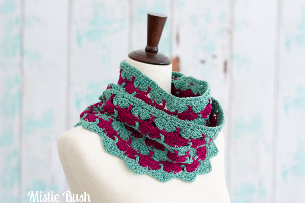 A close-up of the Magade Cowl, a crocheted accessory featuring alternating vertical stripes of jade green and magenta. The cowl displays a lacy, textured stitch pattern and is draped around the neck of a neutral-colored dress form.