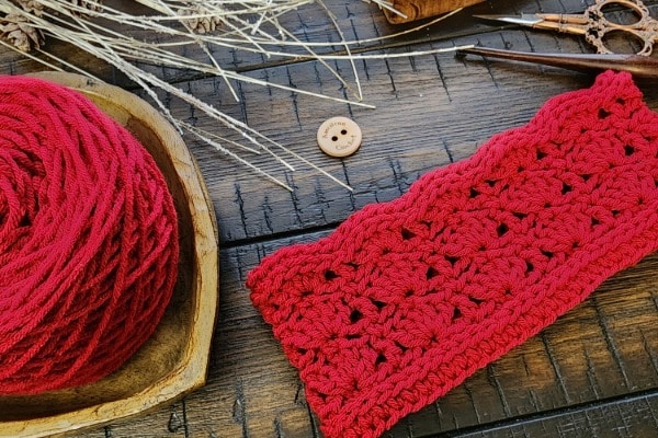 Red textured crochet earwarmer displayed on a wooden surface with matching yarn in a wooden bowl, a ‘Classic’ wooden button, vintage scissors, and rustic dried botanicals.