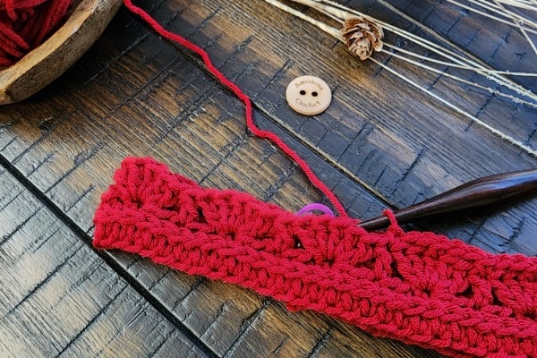 Red crochet earwarmer in progress on a wooden surface with a wooden crochet hook, matching yarn in a bowl, an American Crochet wooden button, and rustic pinecone accents.