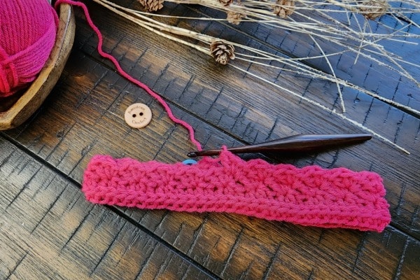 Flat lay of the pink Everyday Kisses Earwarmer on a wooden surface, surrounded by matching yarn, ornate scissors, a crochet hook, stitch markers in a heart‑shaped dish, and a handmade wooden button. Text on the image reads ‘Everyday Kisses Earwarmer – Quick & Easy – Free Crochet Pattern by Mistie Bush, American Crochet.