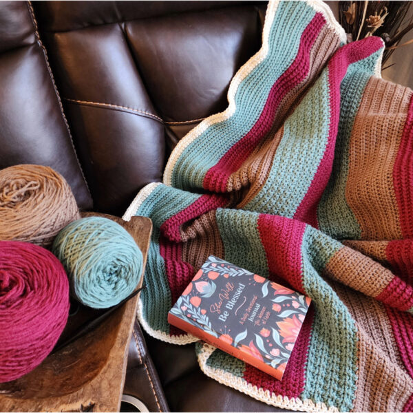 Striped crochet lapghan in teal, burgundy, tan, and cream draped over a brown leather couch, with matching yarn balls in a wooden bowl and a devotional book resting on the blanket.
