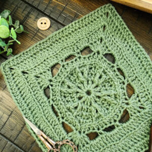 The Faithful Square crochet pattern displayed on a rustic wooden surface with a crochet hook, vintage scissors, and greenery, showcasing its intricate stitch design.