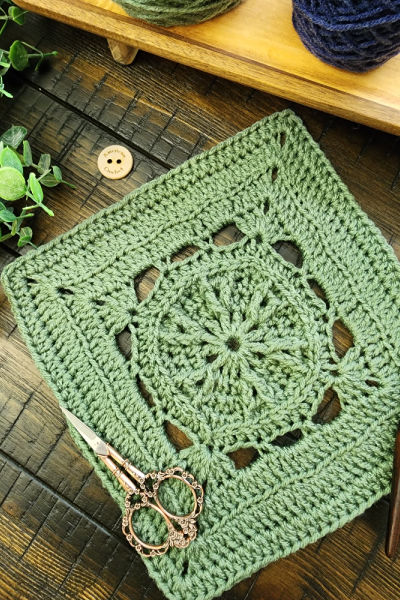 The Faithful Square crochet pattern displayed on a rustic wooden surface with a crochet hook, vintage scissors, and greenery, showcasing its intricate stitch design.