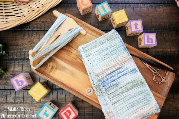 Handmade crochet burp cloth in soft neutral tones, displayed on a wooden tray with crafting tools.
