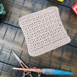 A beige crochet bonding square made using the herringbone single crochet stitch, displayed with a wooden crochet hook, ornate scissors, and baby blocks on a dark wooden surface.