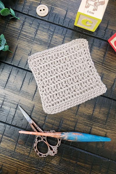 A beige crochet bonding square made using the herringbone single crochet stitch, displayed with a wooden crochet hook, ornate scissors, and baby blocks on a dark wooden surface.