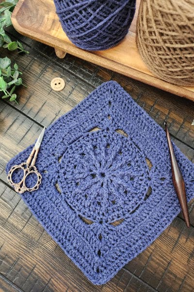 Backroads Square crochet block styled with a wooden crochet hook and ornate scissors, showcasing the stitch textures against a warm wooden background.