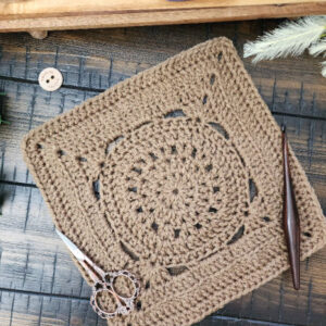 The Pilot Square crochet pattern displayed on a rustic wooden surface with a crochet hook, vintage scissors, and greenery, showcasing its intricate stitch design.